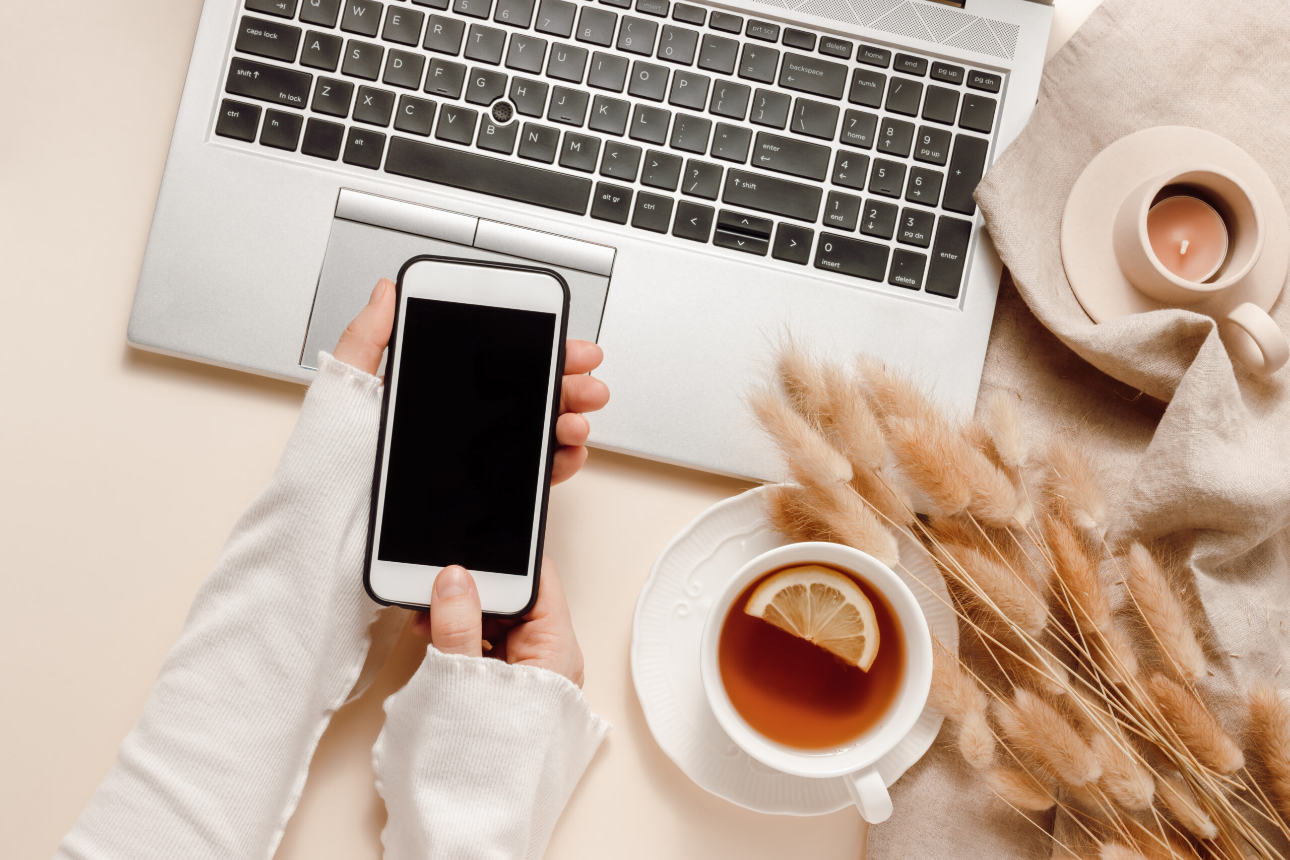 Woman hands hold mobile phone at laptop background with tea, can